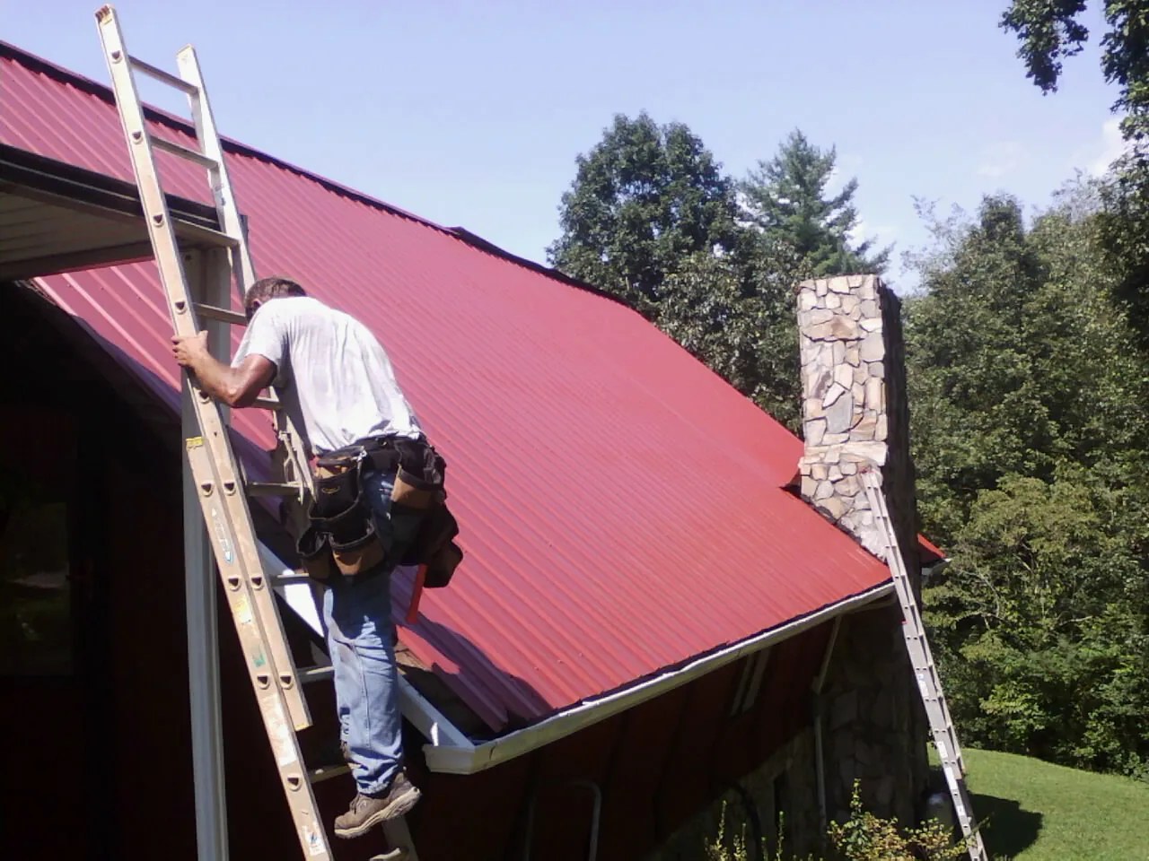 Roofer on a ladder beside a red metal roof and stone chimney — Soffit & Fascia Repair by Roofing Brothers in Willowbrook